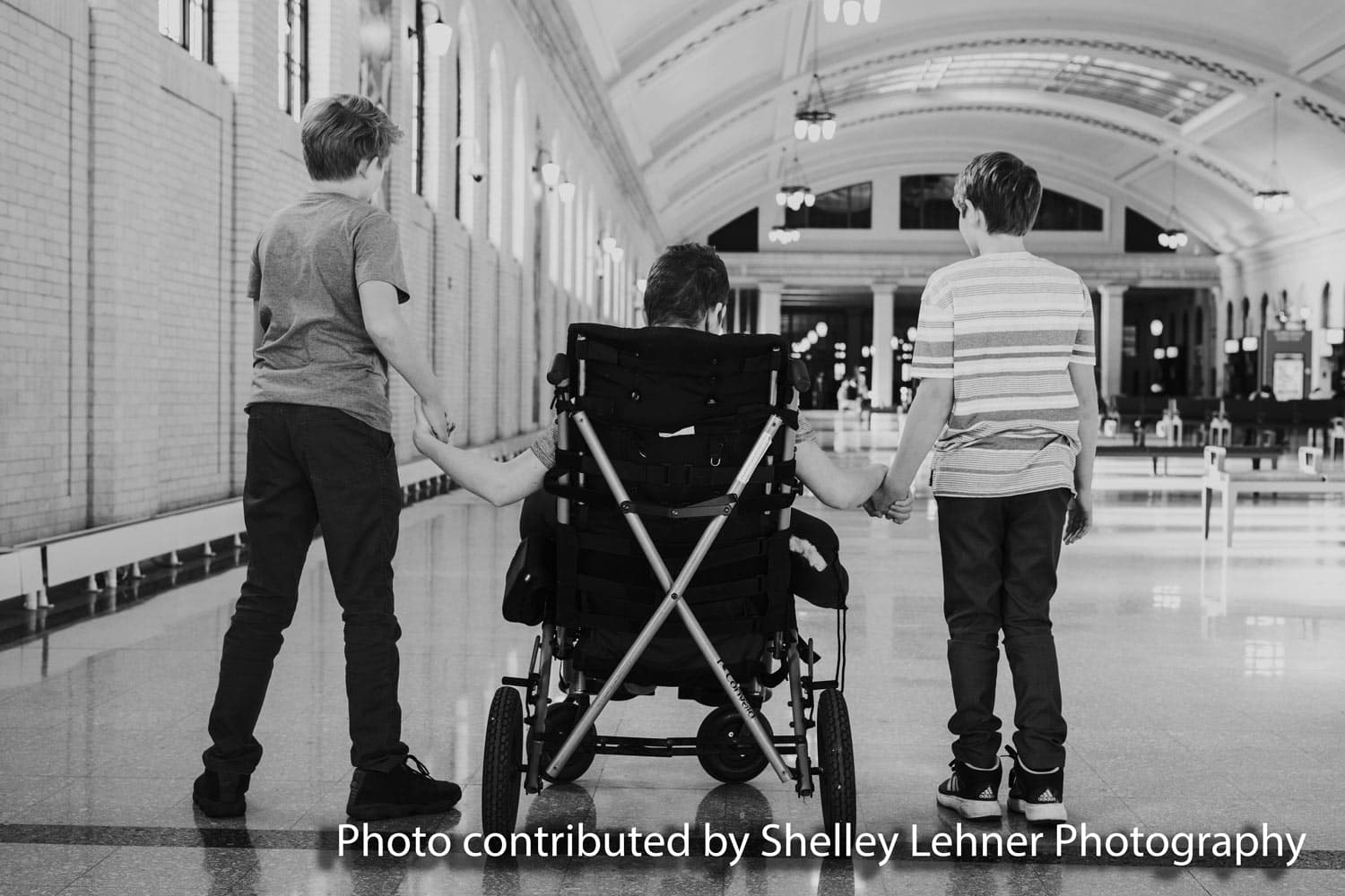 Black-and-white photograph of three brothers seen from behind in a grand station hall, the two standing brothers each holding a hand of their brother seated in his wheelchair
