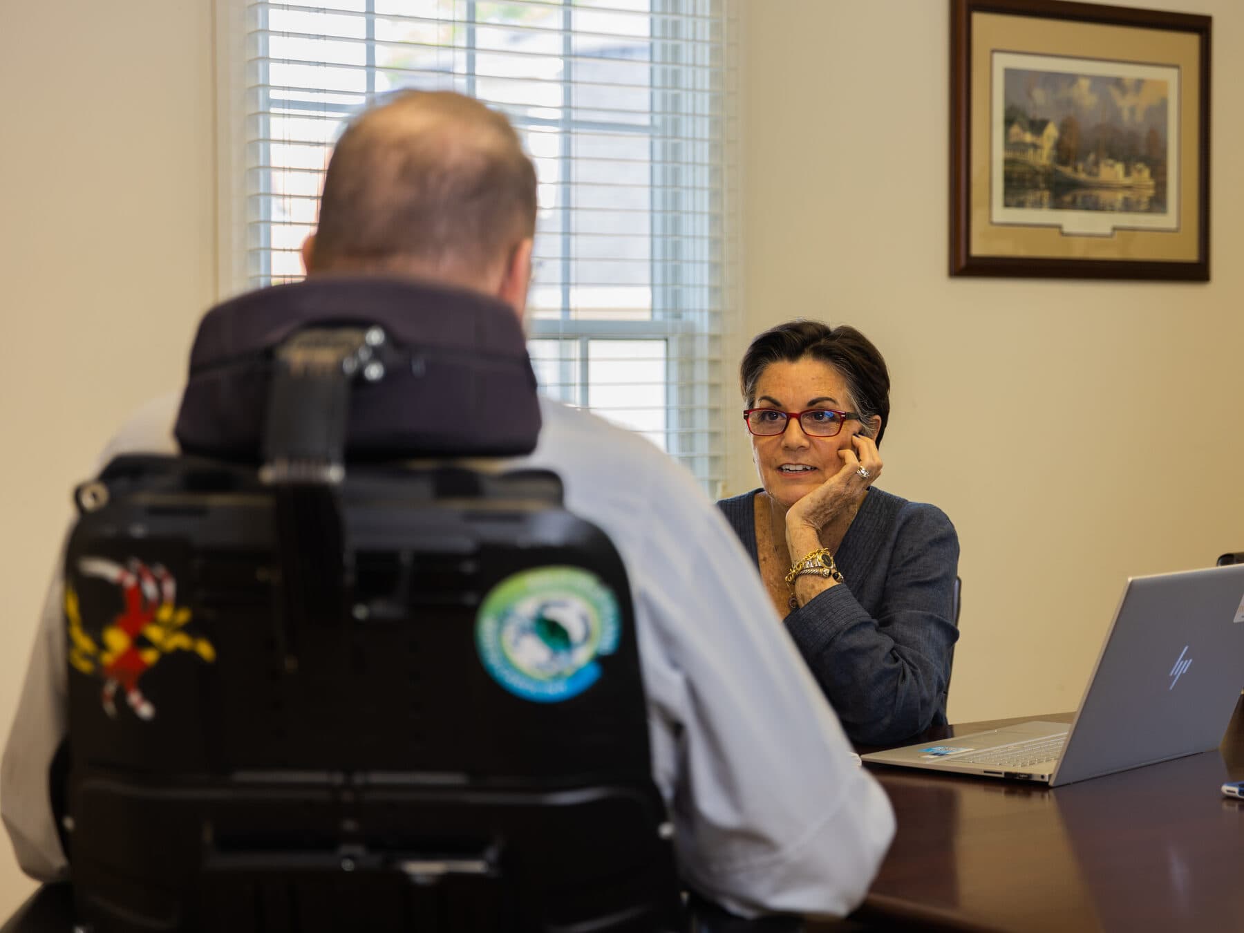 Leigh Ann Kingsbury sitting at a table, smiling in conversation with a man seated in his power wheelchair, seen from behind