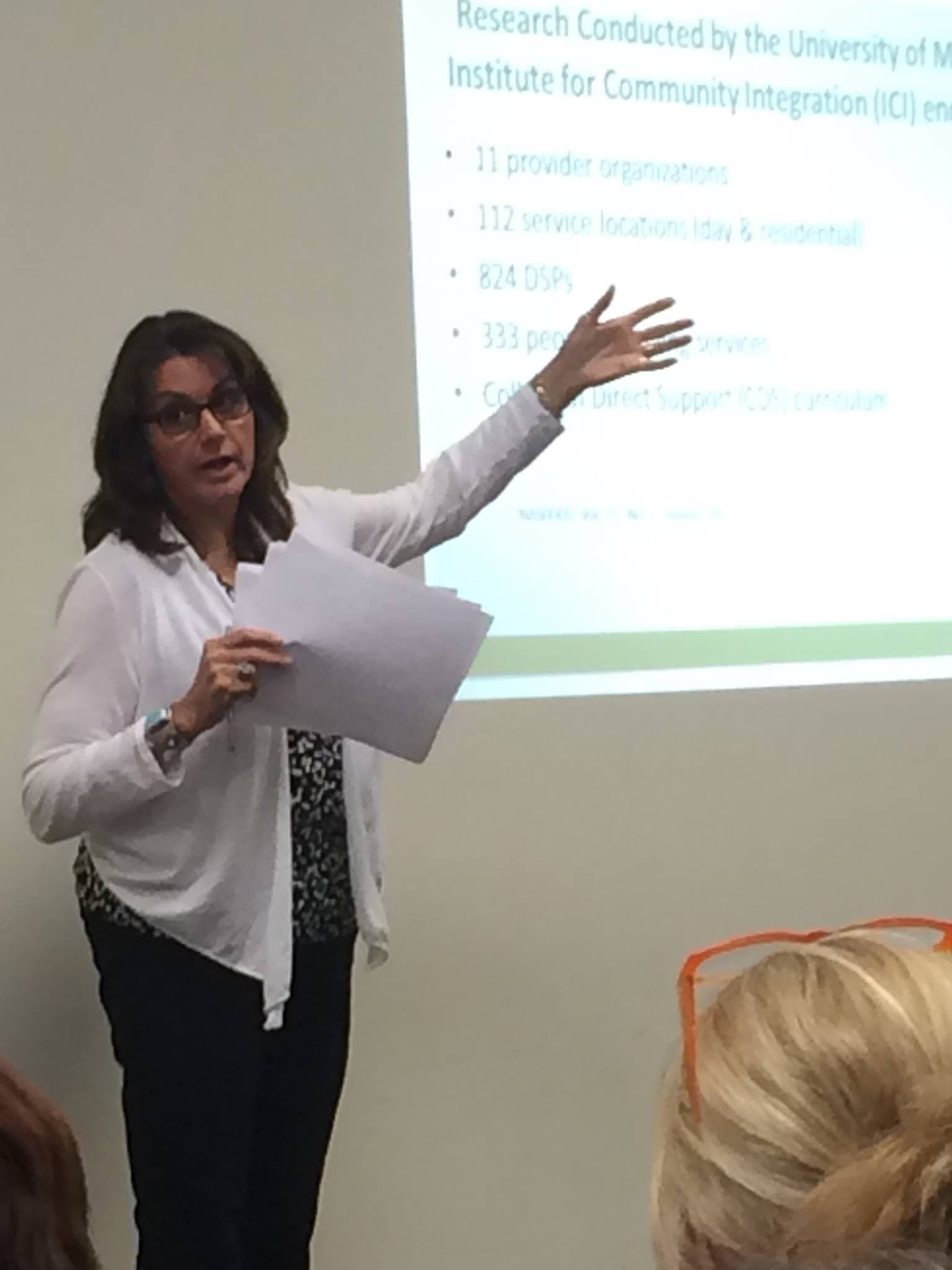 Leigh Ann Kingsbury standing in front of a projected slide of research findings, gesturing to the screen while talking to a seated group